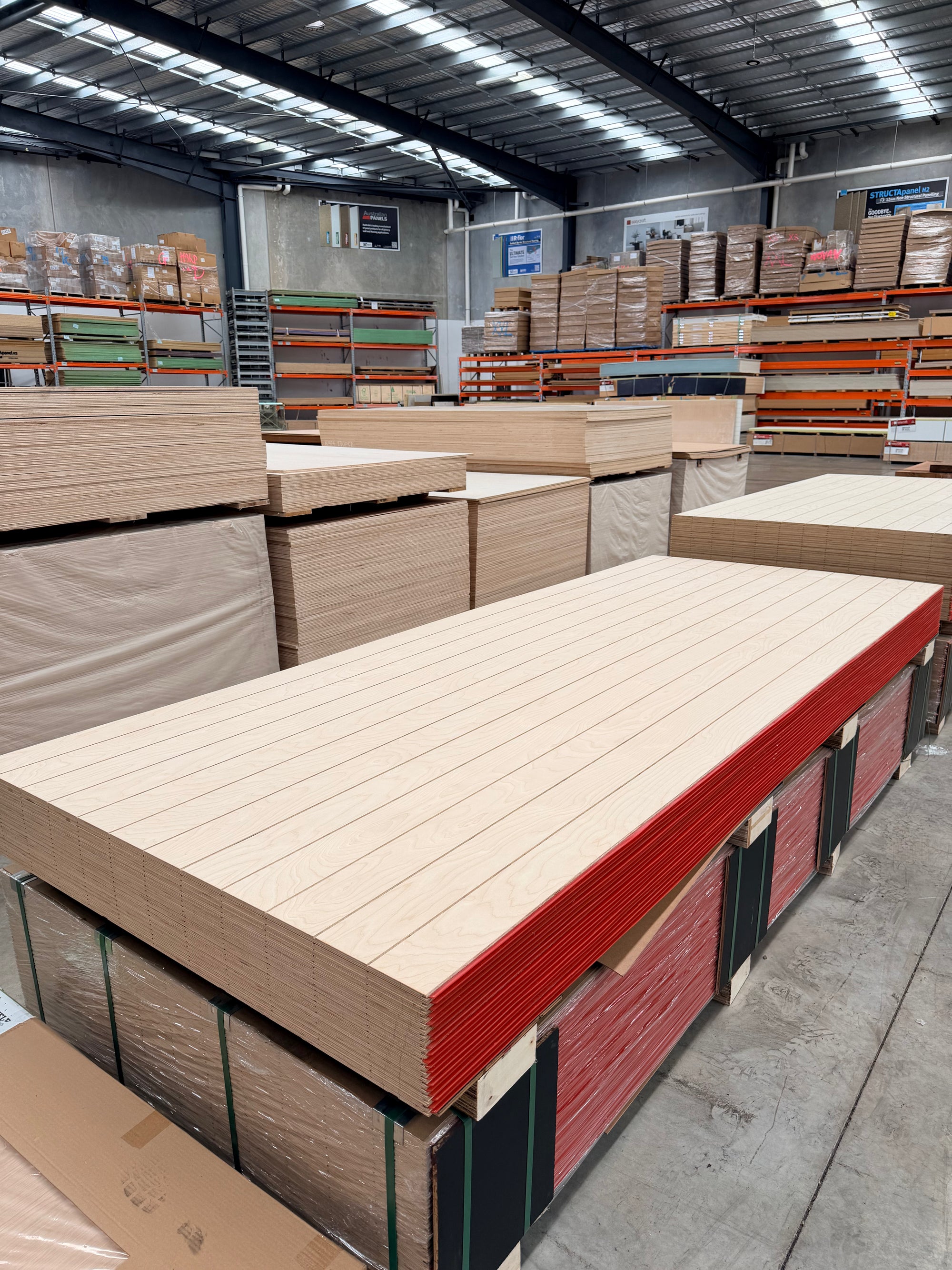 Stack of grooved plywood sheets in a warehouse with shelving and pallet storage
