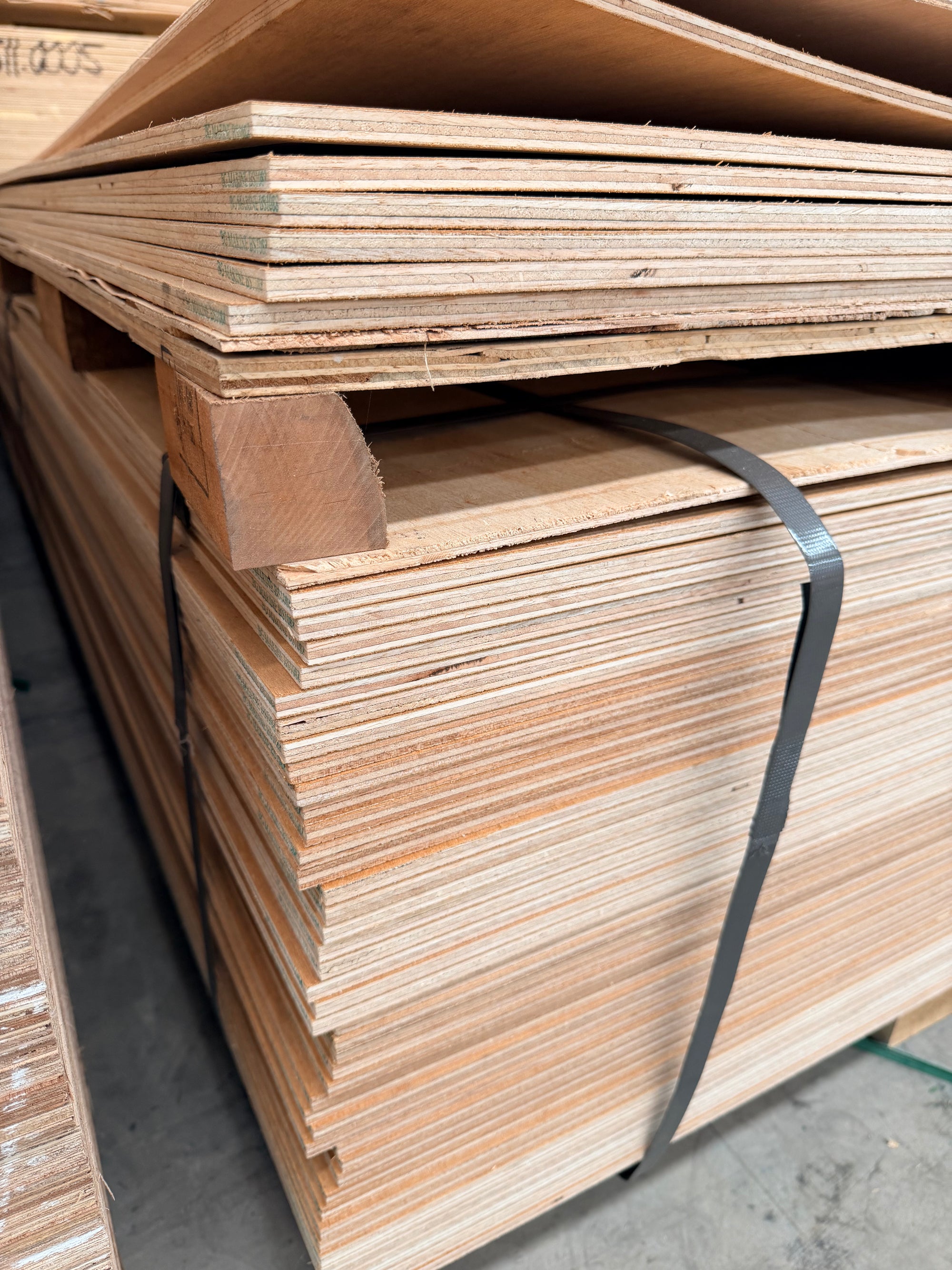 Stack of plywood sheets on a pallet in a warehouse, secured with black straps